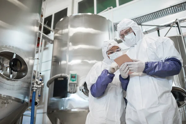 depositphotos_463336922-stock-photo-low-angle-view-two-workers Infant formula consulting experts in maximum-level safety suits using a tablet to inspect large processing tanks in a dairy facility.